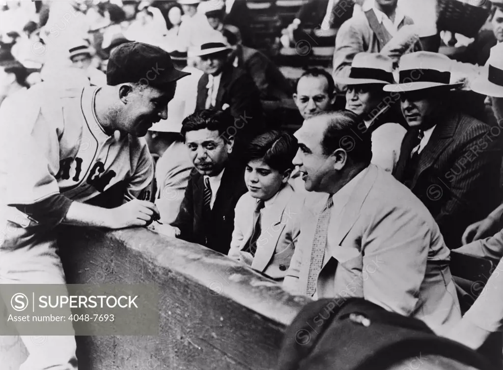 Chicago Cubs player Gabby Hartnett autographing a baseball for Sonny Capone who is sitting with his father Al Capone and other gangsters at a 1931 charity baseball game.