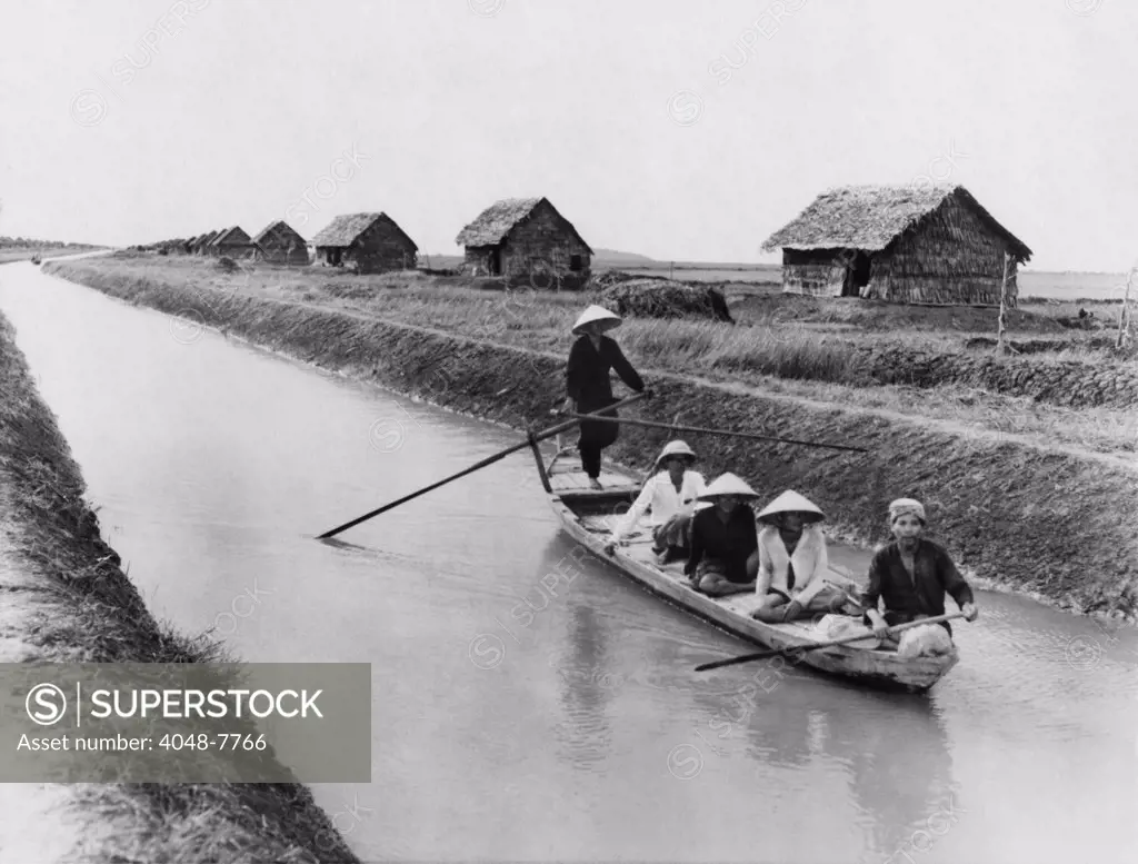 Vietnamese men in a sampan on a canal near straw dwellings that house refugees from North Vietnam. 1956. LC-DIG-ppmsca-09168