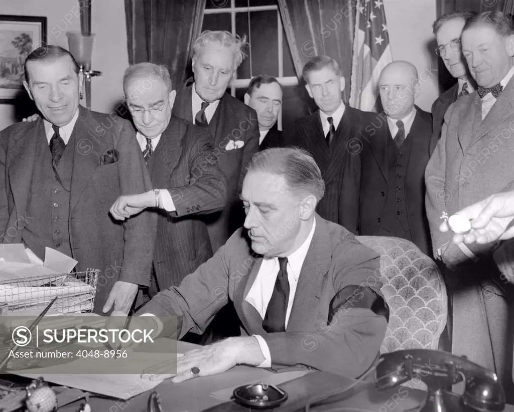 President Franklin D. Roosevelt signs the joint congressional resolution declaring war with Japan at 4:10 PM, Dec. 8, 1941. Standing L-R: Rep. Sol Bloom, Luther Johnson, timing the signing, Charles Eaton , Joseph Martin, Vice President Henry Wallace, Speaker Sam Rayburn, John McCormack, Massachusetts, and Senator Charles McNary. FDR wears a black arm in mourning for his recently deceased mother, Sara Delano Roosevelt.