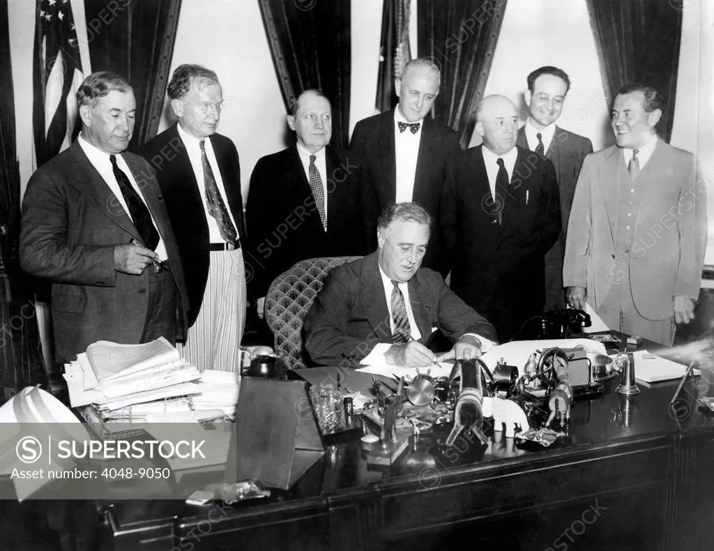 President Franklin Roosevelt, signs the Public Utility Holding Company Act of 1935 (Wheeler-Rayburn Act). It imposed consumer protection regulations on public utilities and was bitterly fought by the industry. Standing L-R: Sen. Alben Barkley, Sen. Burton Wheeler, Sen. Fred H. Brown, Dozier Du Vane, Solicitor, Federal Power Commission, Rep. Sam Rayburn, Benjamin Cohen, Attorney, and Thomas Corcoran, attorney. Aug. 26, 1935.