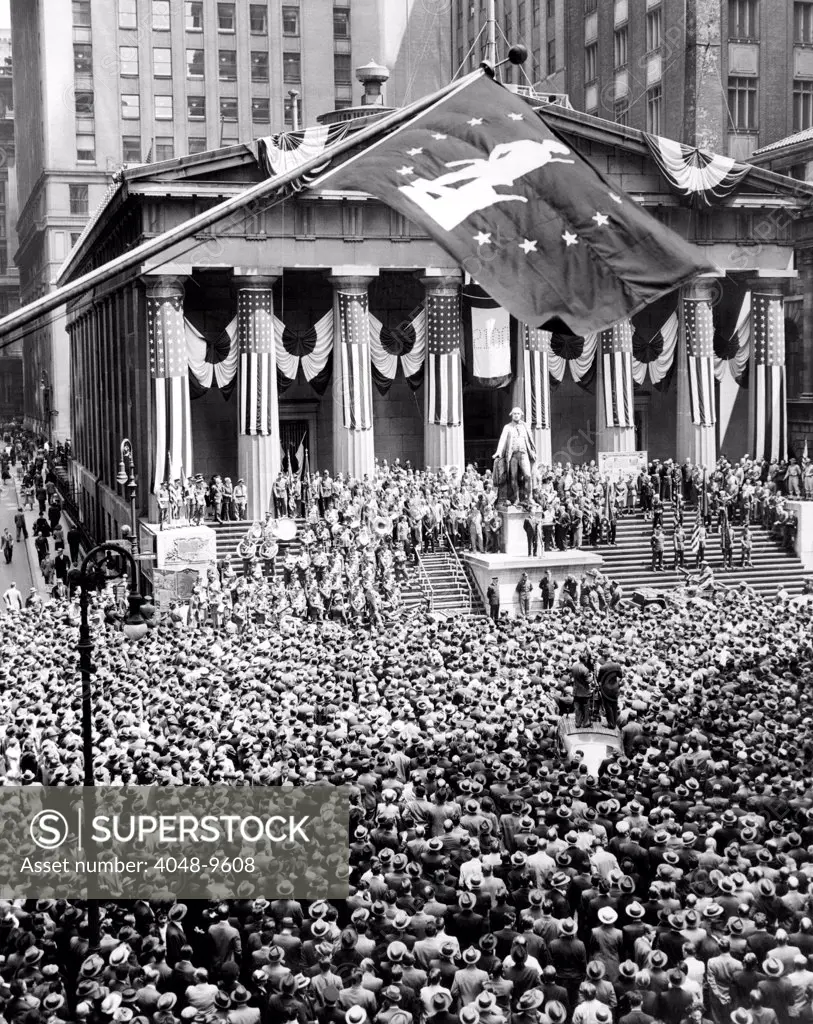 The New York Stock Exchange celebrates its 150th anniversary with the greatest war bond rally in the city's history. The war bond flag waves above the statue of George Washington at the Subtreasury Building. May 8, 1942.