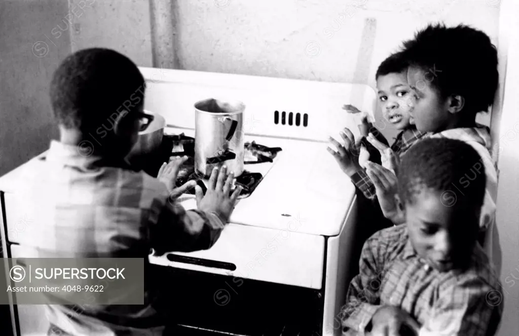 Four African American children cluster around the gas stove for warmth. The inadequate heating in their Harlem apartment encourages use of the dangerous alternative when heat is not provided by slum landlords. April 6, 1968.