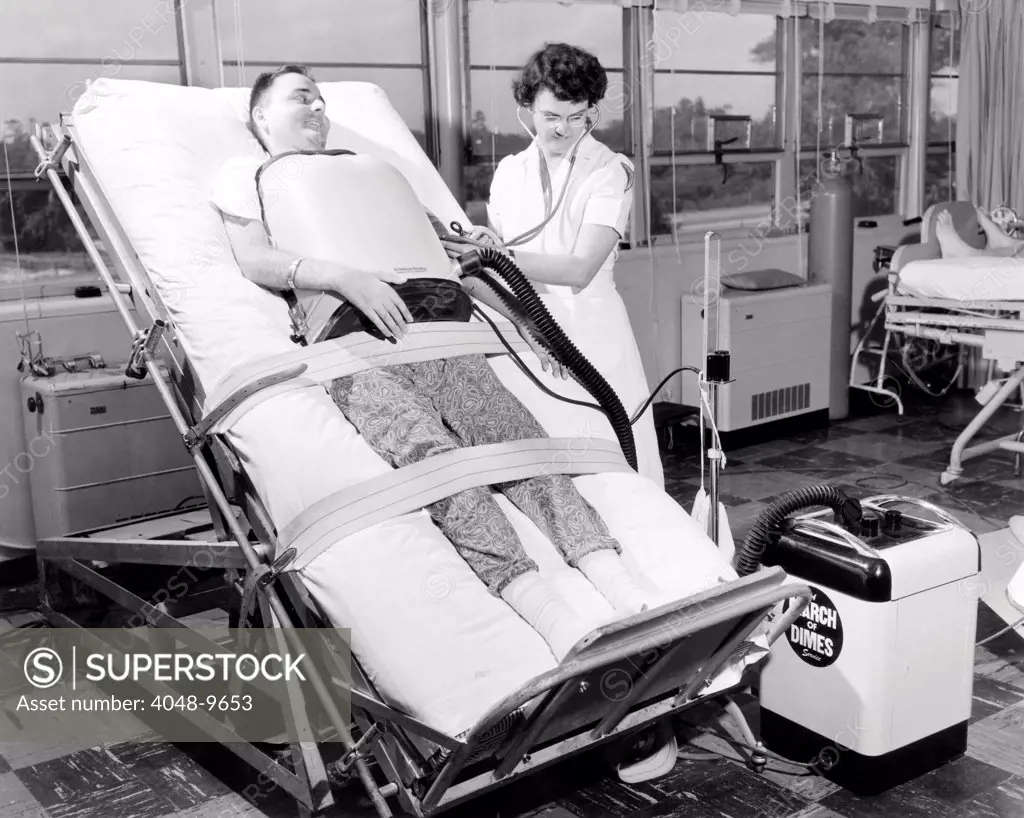 A 30 year old polio patient wearing a chest respirator on a standing bed. This relieves pressure on the back from the horizontal bed of iron lungs. The nurse checks the patient's blood pressure as he moves into a vertical position. Jefferson Davis Hospital, Houston, Texas. 1953.