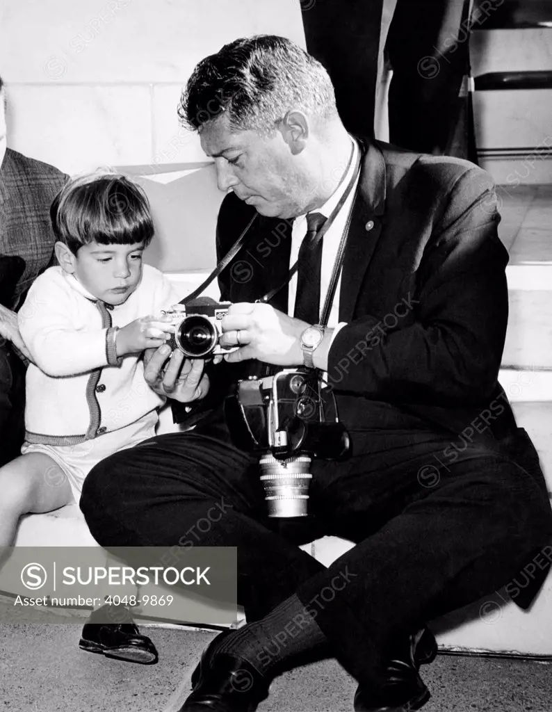 John F. Kennedy Jr. takes an interest in the cameras of White House photographer Capt. Cecil Stoughton. Nov. 11, 1963.