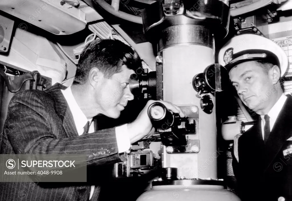 President John Kennedy looks through the periscope of the nuclear submarine USS Thomas A. Edison. At right is his Naval Aide, Capt. Tazewell Shepard.