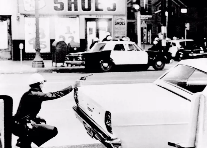 1965 Watts Riots. A policeman aims his revolver at building where a sniper shot at passing cars. Across the street, fellow officers crouch behind cars as they move in closer. Moments later, National Guardsmen returned the snipers fire, and thought they had hit him. August 14, 1965.