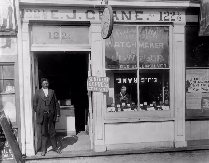 African American watchmaker, original title: 'E.J. Crane, watchmaker and jewelry store with man working in window and man standing in doorway', Richmond, Virginia, photograph, 1899.