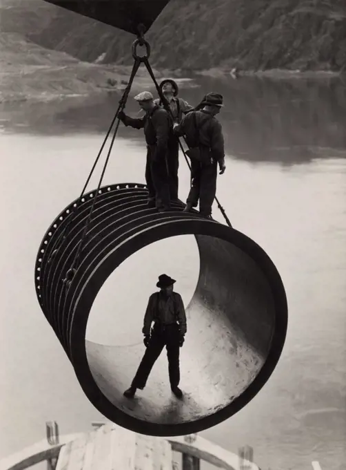 Four workers riding on a large casing section of pipe suspended from cable as it is moved into position during construction (1933-42) of the Grand Coulee Dam, Washington.