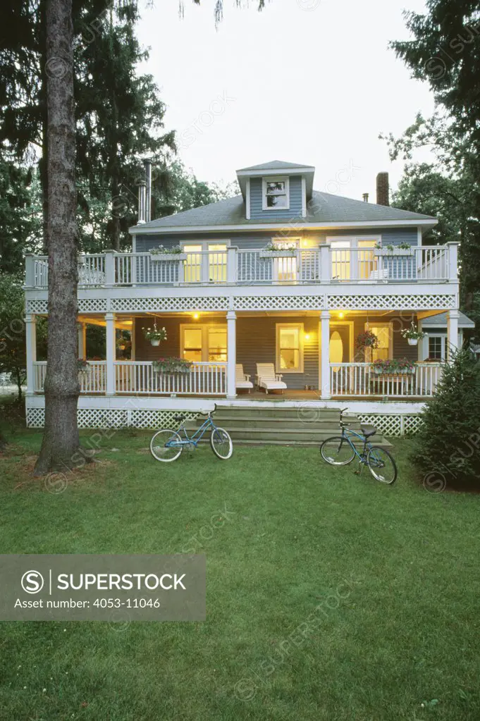 EXTERIORS: Two-story single family four square farmhouse house, wraparound porch on two levels, flower boxes on railings, hanging flowers, third floor dormer, bicycles on lawn, twilight, vertical