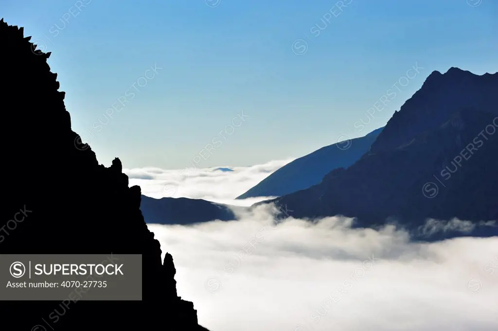View over silhouetted jagged mountain edge and mountains covered in mist at sunrise seen from the Col du Tourmalet, Pyrenees, France, June 2012