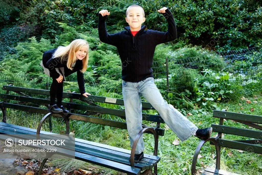 Young Boy on Park Bench Flexing Arms with Young Girl Smiling Behind Him
