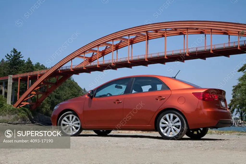 Rear 3/4 view of a 2010 red Kia Forte in front of an arch bridge