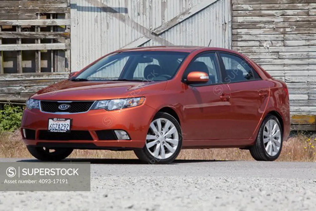 Front 3/4 static view of a 2010 red Kia Forte on a rural road in front of an old barn
