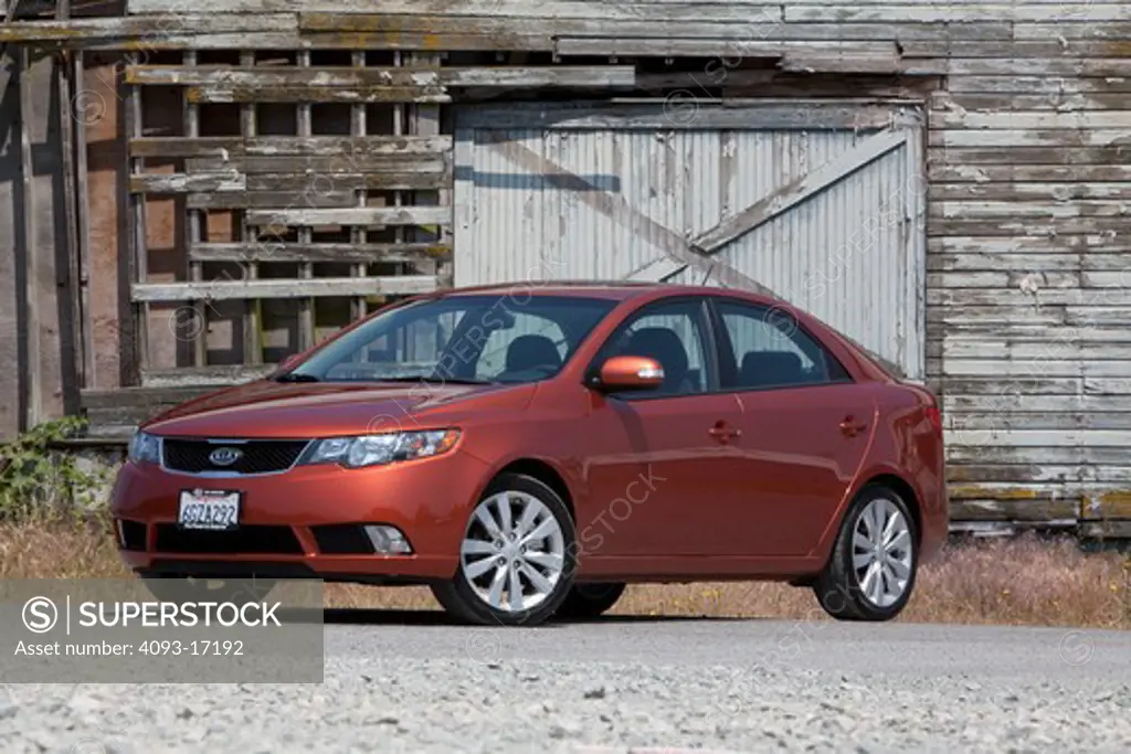 Front 3/4 static view of a 2010 red Kia Forte on a rural road in front of an old barn