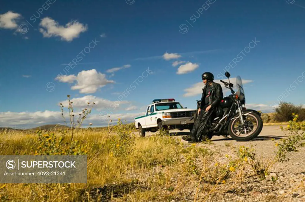 man biker stopped and leaning standing next to a motorcycle harley while parked in front of a truck with lights park ranger looking out.