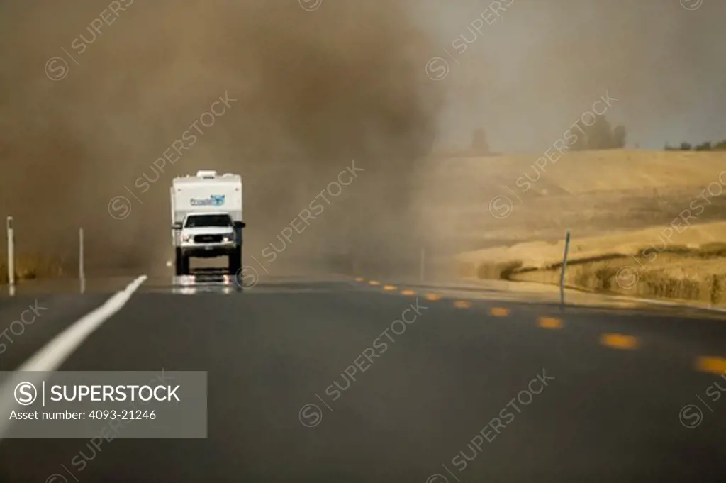 front nose view of a truck towing a camper in front of a large dust storm in an open field.