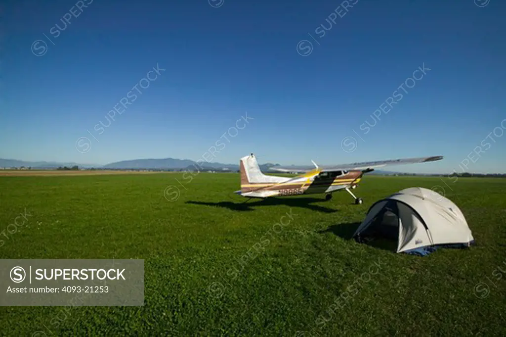 airplane stopped in green wide field next to a camp site tent. rear 3/4 view.