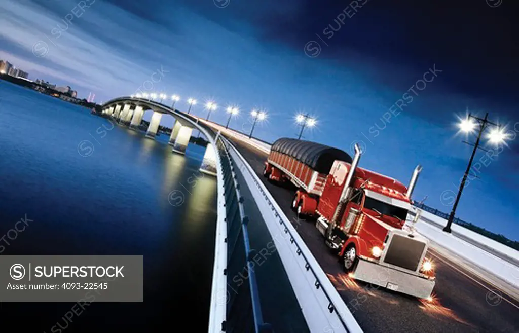 A red Peterbuilt demi and covered load trailer crossing a bridge at dusk.