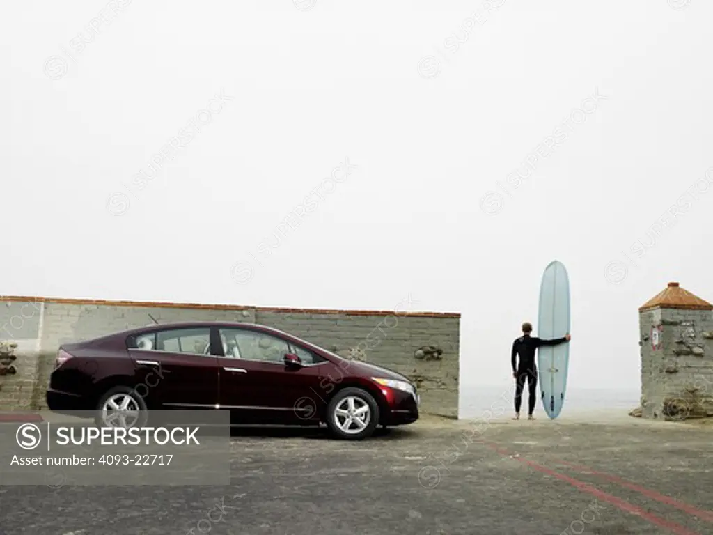 Profile static view of a 2009 Honda FCX Clarity, hydrogen fuel-cell ( fuel cell ) powered vehicle. Surfer holding a surfboard in the background.
