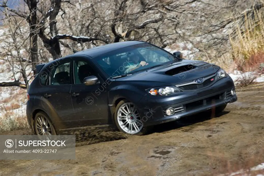 Front 3/4 action view of a 2009 Subaru Impreza WRX STi on a muddy, rural road kicking up mud.