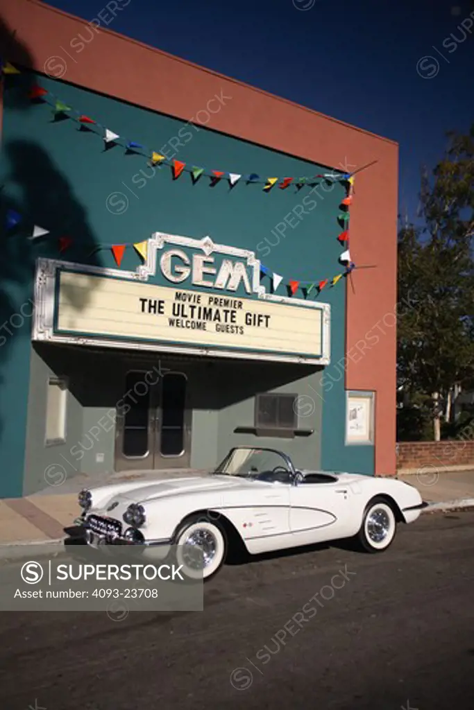 Front 3/4 static view of a white 1958 Chevrolet Corvette in front of a classic movie theater. Taking its name from the corvette, a small, maneuverable fighting frigate, the first Corvettes were virtually handbuilt. The outer body was made out of a revolutionary (at the time) new composite material called fiberglass. Underneath that radical new body were standard Chevrolet components, including the Blue Flame inline six-cylinder engine, two-speed Powerglide automatic transmission, and drum brakes