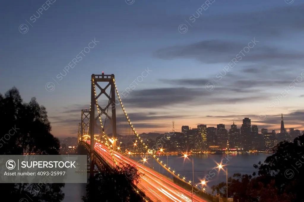 dusk shot of the bay bridge lit up with passing cars leaving light streaks across and the downtown buildings in view.