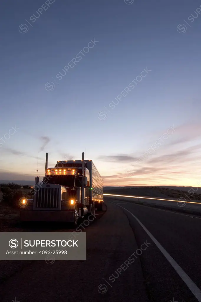 A big Rig pulled over and stopped on the side of a freeway with his parking lights on and light streaks from passing cars in the background during dusk.