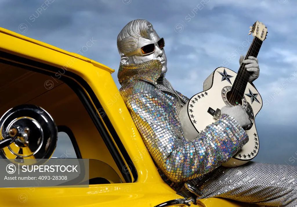 Silver Elvis impersonator, street performer with a guitar sitting on the side of a classic yellow 1963 Chevrolet C10 fleetside pickup truck