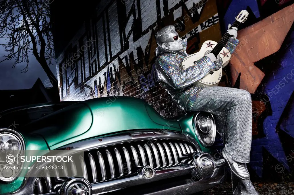 Silver Elvis impersonator, street performer with a guitar and sitting on the hood of a 1953 Buick Roadmaster Riviera Coupe and graffiti painted street