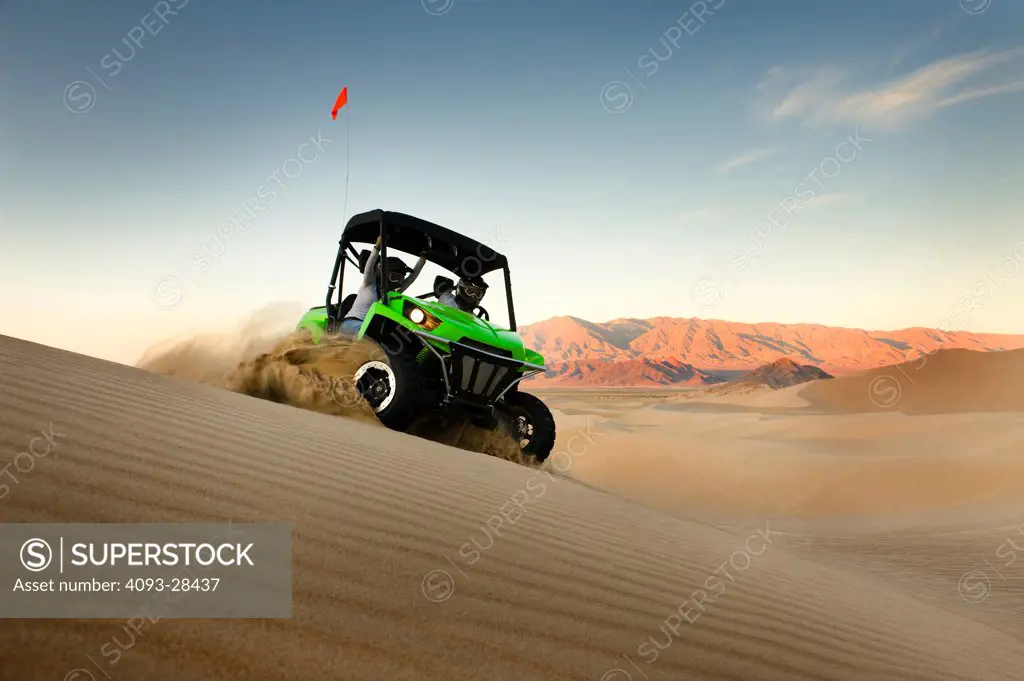 Front 3/4 action of a green 2010 Kawasaki Teryx Quad ATV kicking up sand on a large sand dune at dusk