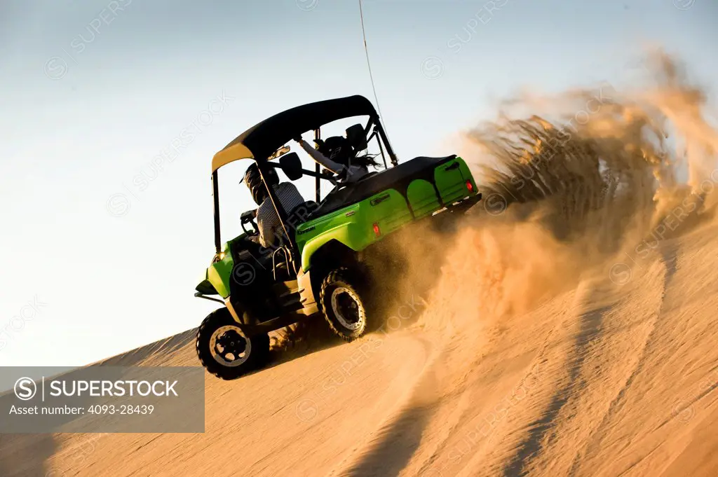 Rear 3/4 action of a green 2010 Kawasaki Teryx Quad ATV kicking up sand on a large sand dune at dusk