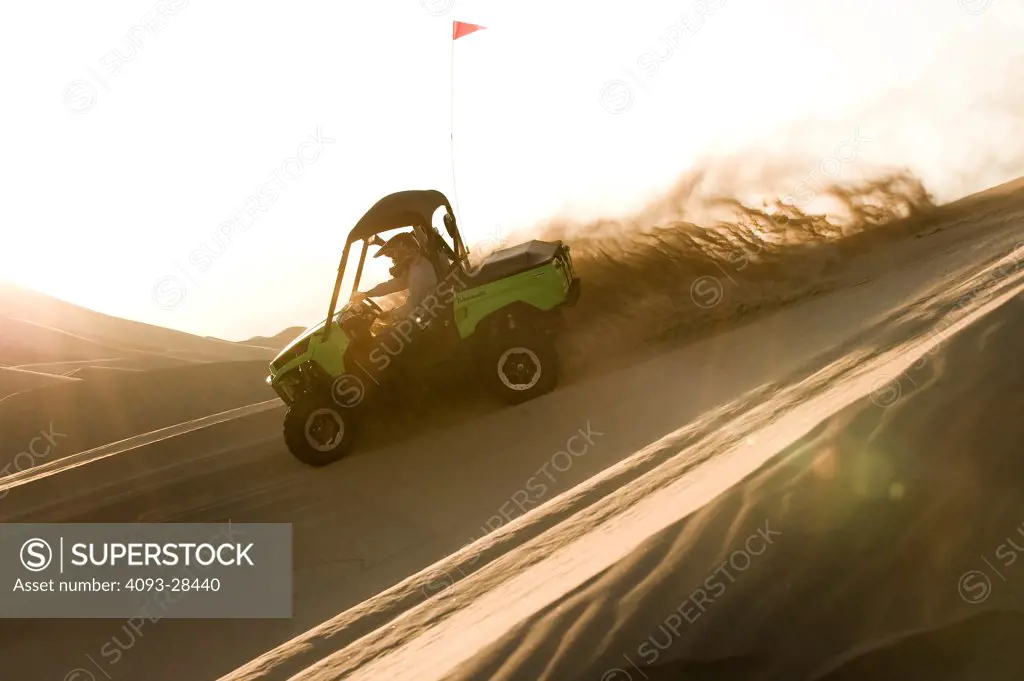 Profile action of a green 2010 Kawasaki Teryx Quad ATV kicking up sand on a large sand dune at dusk