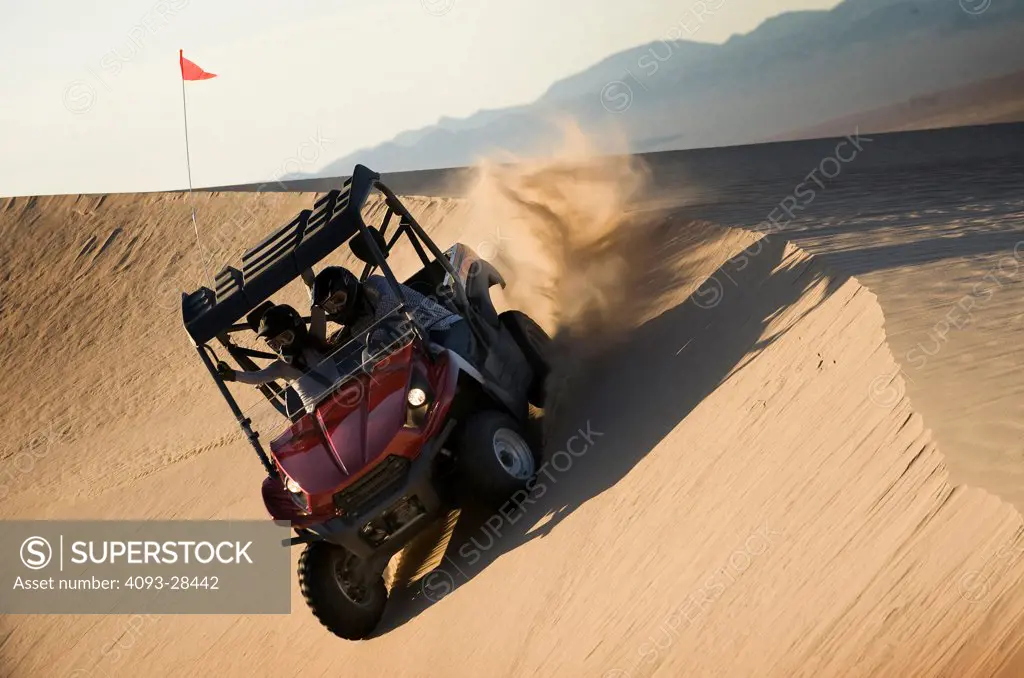 Front 3/4 action of a red 2010 Kawasaki Teryx Quad ATV kicking up sand on a large sand dune in the late afternoon