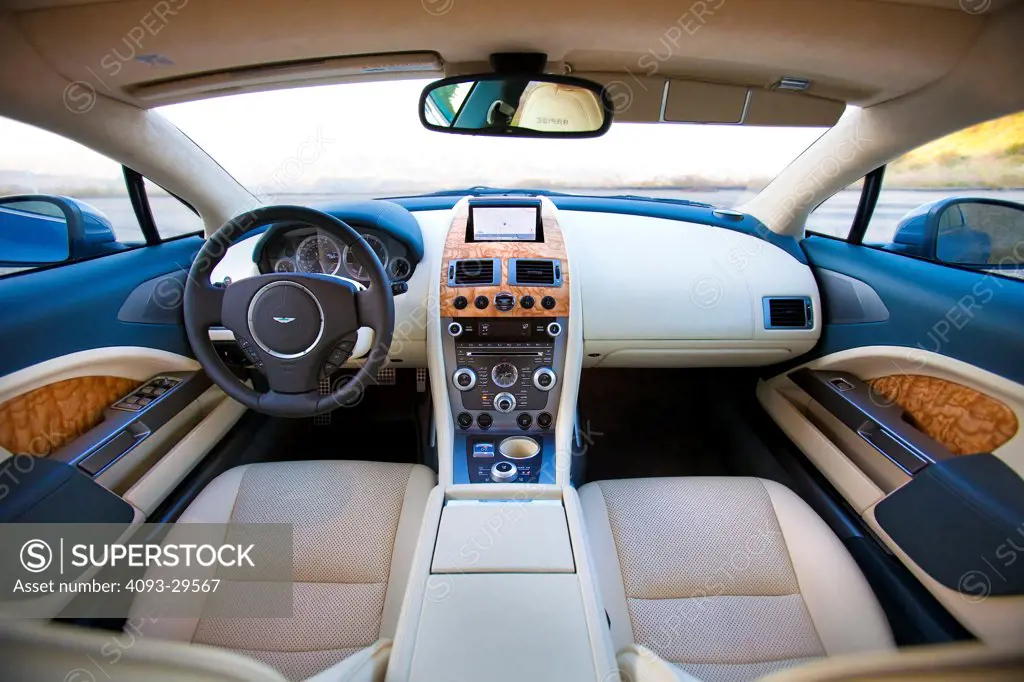 Interior view of a blue 2012 Aston Martin Rapide showing the steering wheel, instrument panel, center console, dashboard and tan leather front seats from the rear.