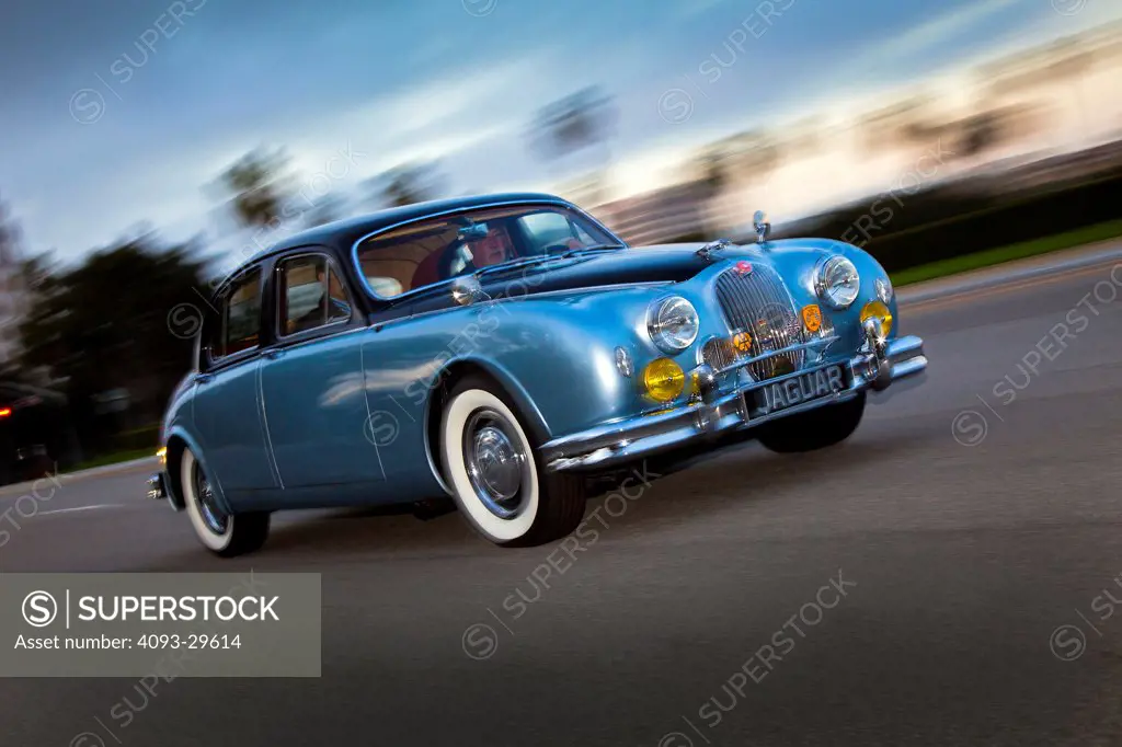 Front 3/4 action of a two tone blue or black 1955 Jaguar Mark 1 on a suburban road at dusk.