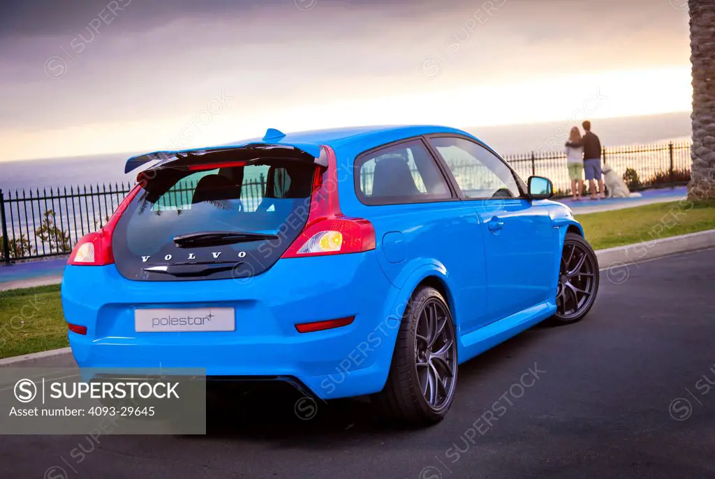 Rear 3/4 static of a blue 2013 Volvo C30 T5 Polestar parked near the ocean at sunset. Couple with dog in the background.