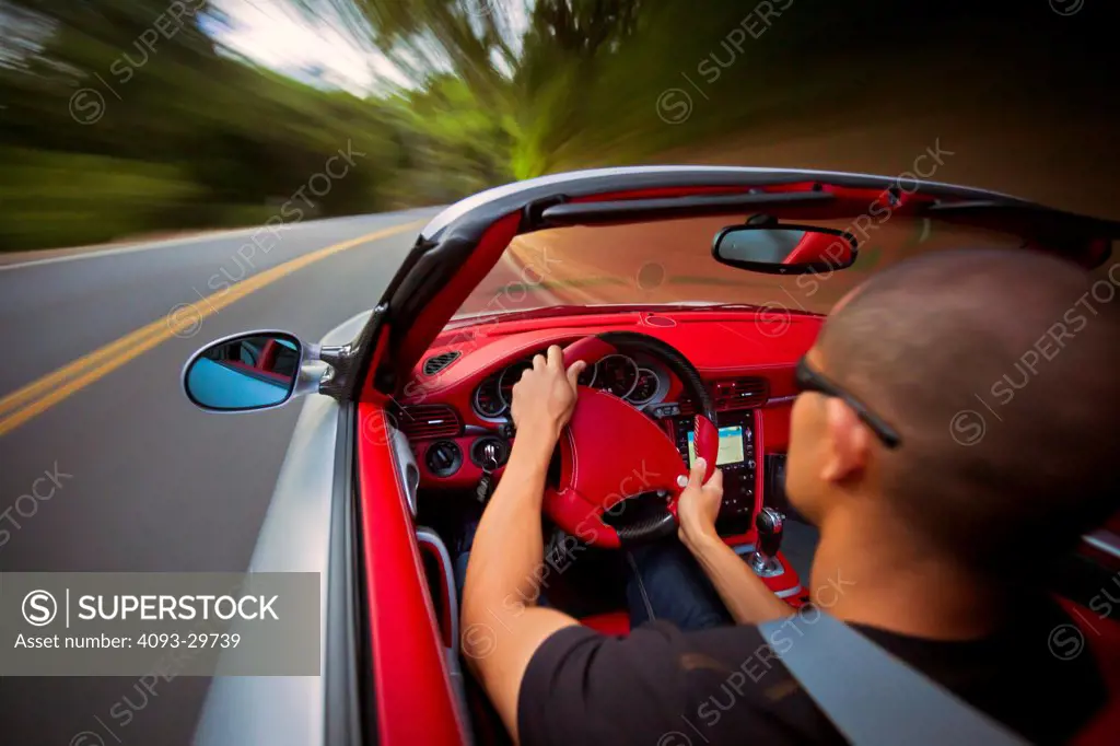 Interior action of a man driving a silver 2013 Ruf RT Roadster with a bright red leather interior. On a rural, tree lined road.