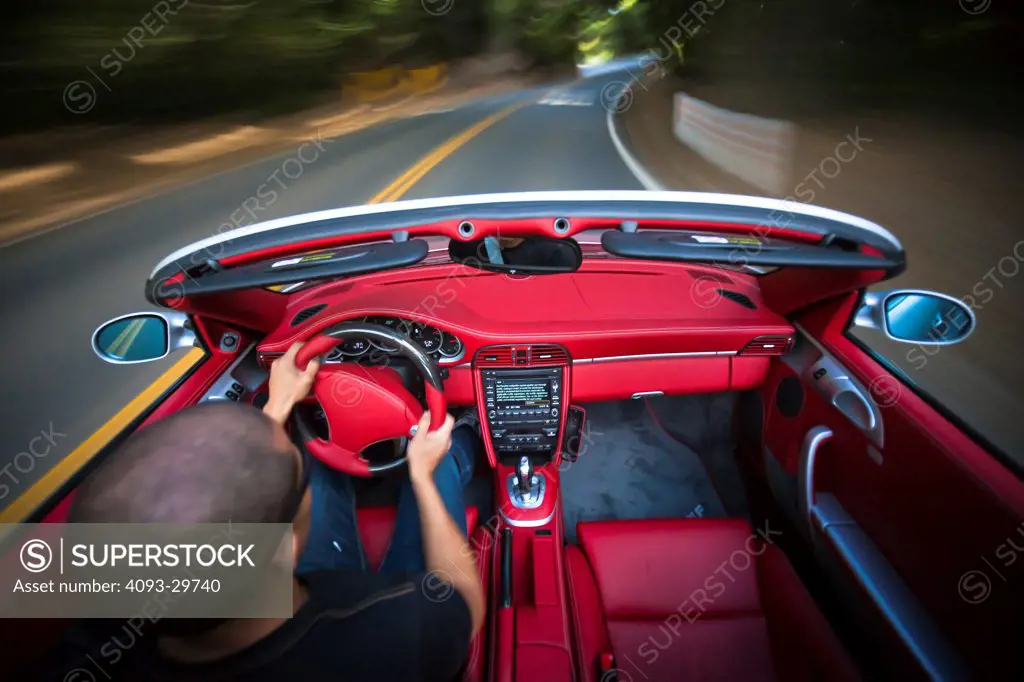 Interior action of a man driving a silver 2013 Ruf RT Roadster with a bright red leather interior. On a rural, tree lined road.