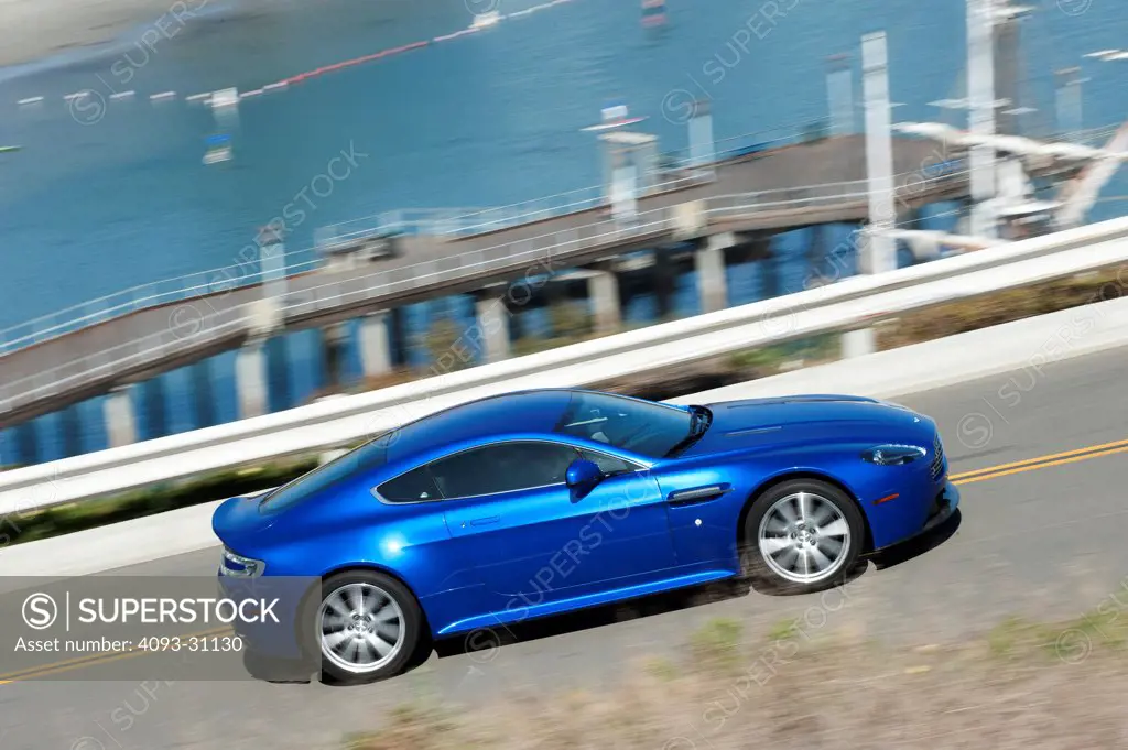 High angle front 7/8 view of a blue 2012 Aston Martin Vantage S driving near a harbor