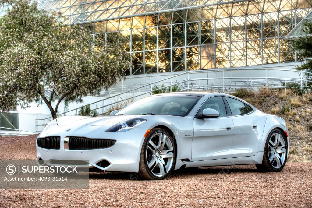 Front 3/4 view of a white 2012 Fisker Karma in front of an unusual house.