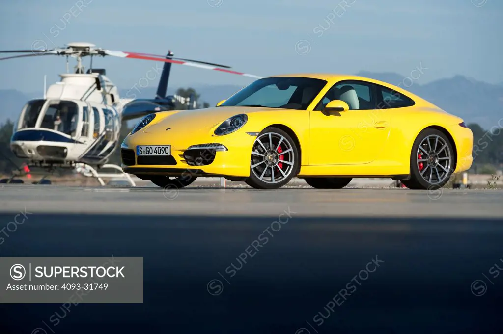 Front 3/4 view of a yellow 2012 Porsche 911 Carrera S, platform number 991, shown with a helicopter in the background.