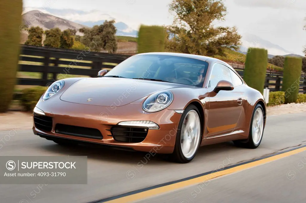 Front 3/4 view of a brown 2012 Porsche 911 Carrera S, platform number 991, driving along a rural road.