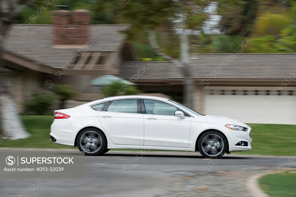 Profile view of a white 2013 Ford Fusion Titanium driving through a suburban neighborhood.