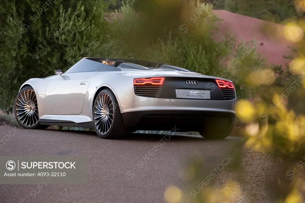 Rear 3/4 view of a convertible silver Audi e-tron Spyder Concept parked in front of a red house in a forested location.