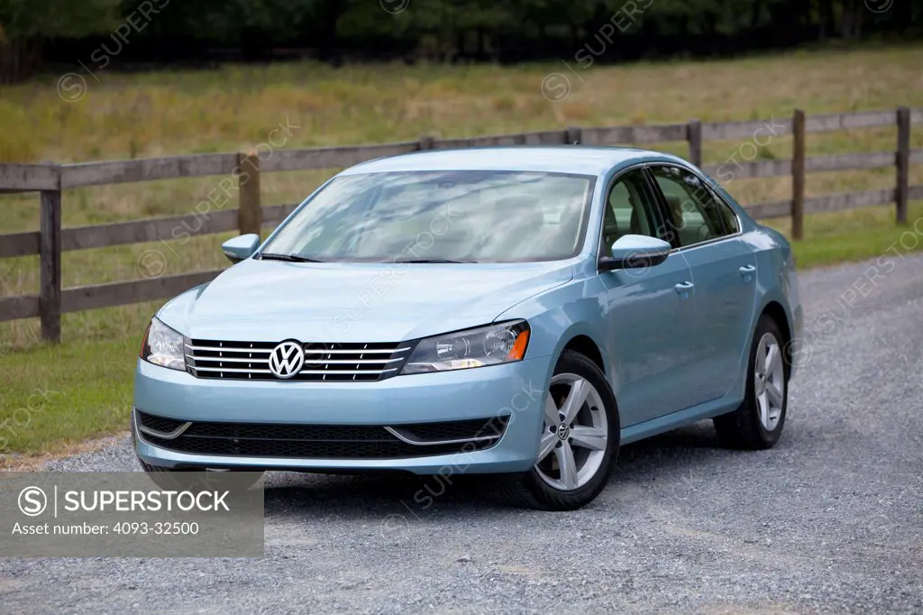 Front 3/4 static view of a light blue 2012 Volkswagen Passat TDI diesel on a rural, gravel road with wood fence in the background.