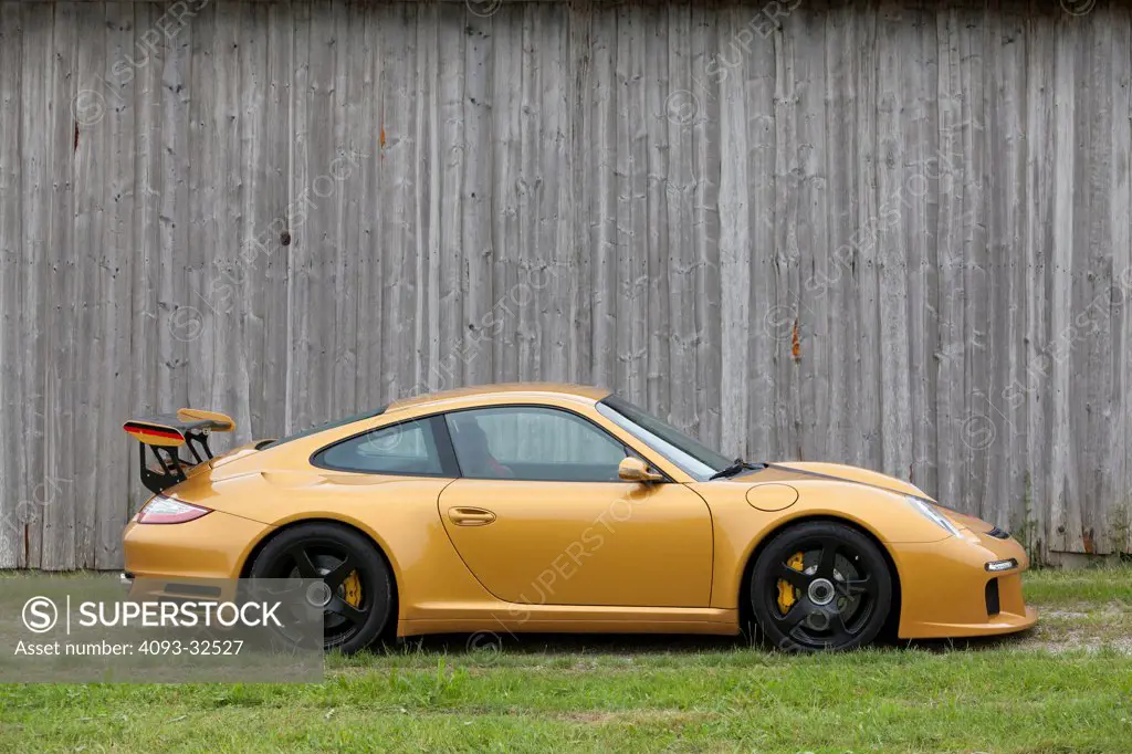 Profile static of a gold 2012 RUF RT12R. Based on a Porsche 911 parked on a gravel road next to an old barn.