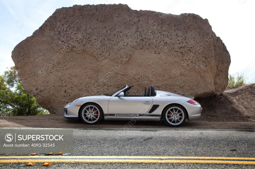 Profile static view of a silver 2012 Porsche Boxster Spyder parked on the side of a rural mountain road and next to a large boulder.