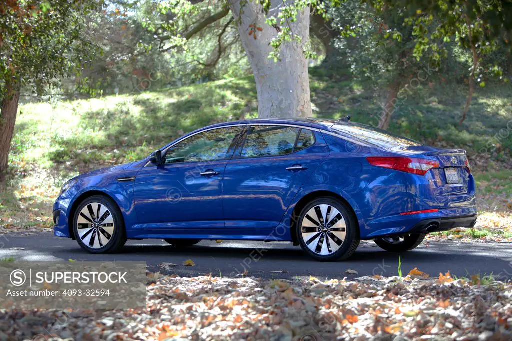 Rear 3/4 static of a blue 2012 Kia Optima on a rural, tree lined road.