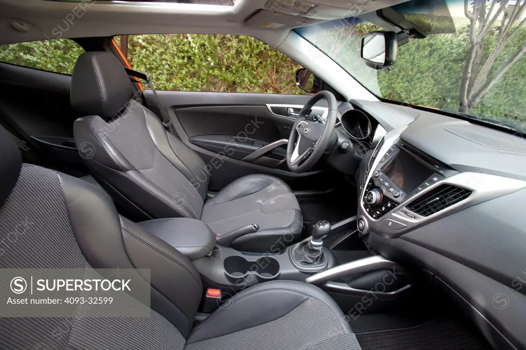 Interior of a 2012 Hyundai Veloster showing the showing the instrument panel, steering wheel, dashboard, center console, multi function display, climate controls, gear shift lever, parking brake and black leather seats.