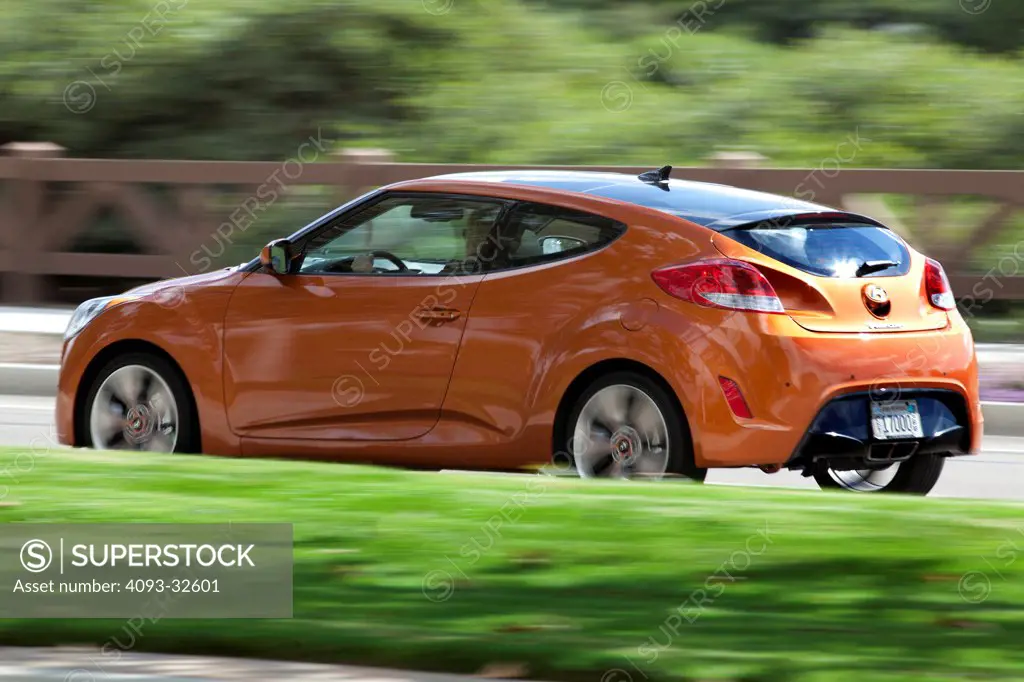 Rear 3/4 action of an orange 2012 Hyundai Veloster on a rural road.